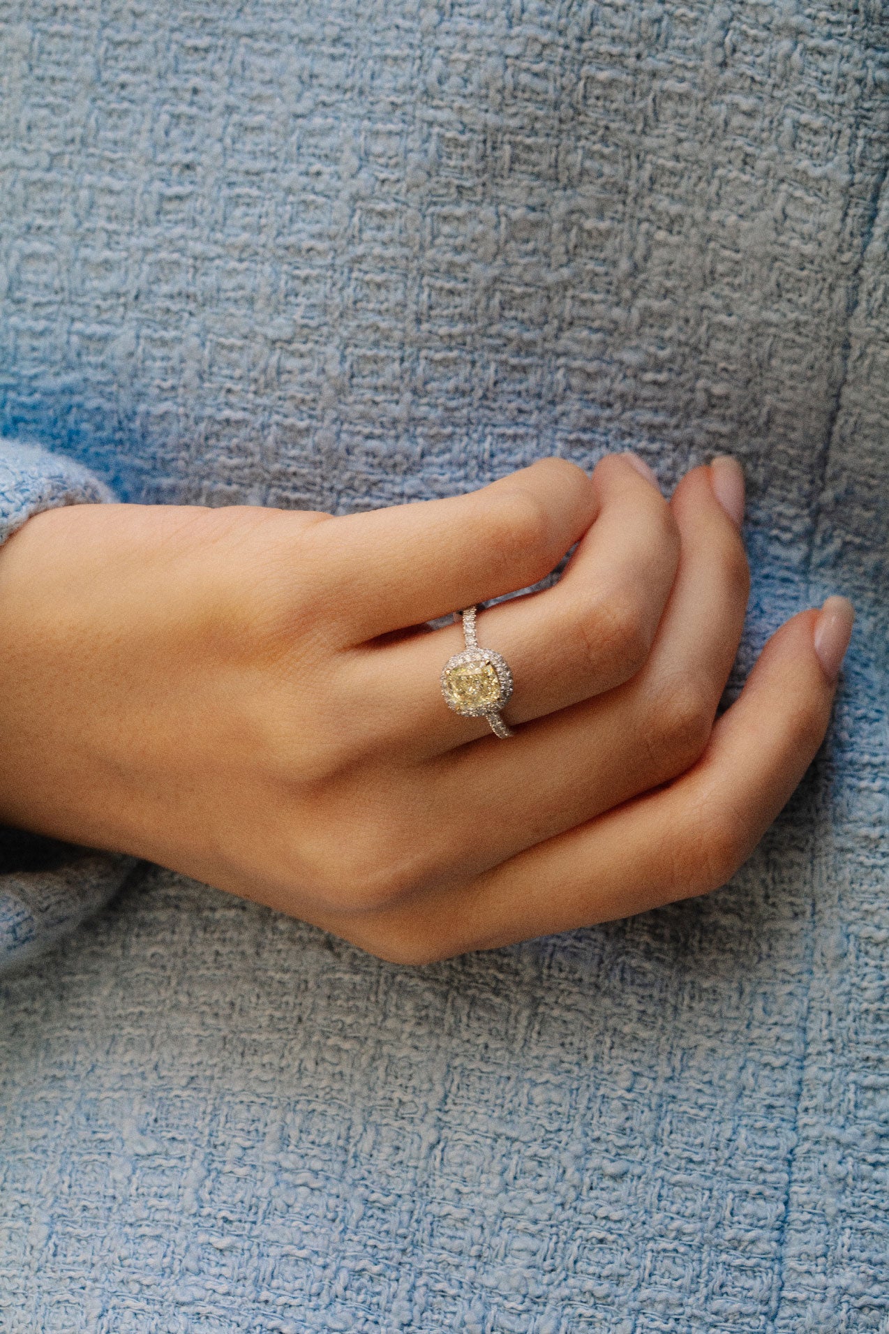 Photo d'une main portant la magnifique bague Santoline en or blanc. Cette bague d'une pierre centrale diamant jaune et d'un halo de diamants blancs, ajoute une touche de sophistication à la tenue de la femme en veste en tweed bleue.