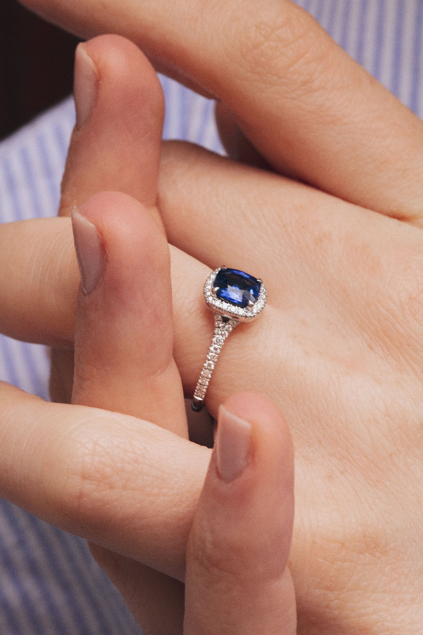 Photo zoomé des mains d'une femme qui s'entrelacent. Une bague en saphir bleu habille son doigt d'un glamour extrême. Le halo de diamants blancs et du pavage sur la monture magnifient l'éclat bleu de la pierre centrale..