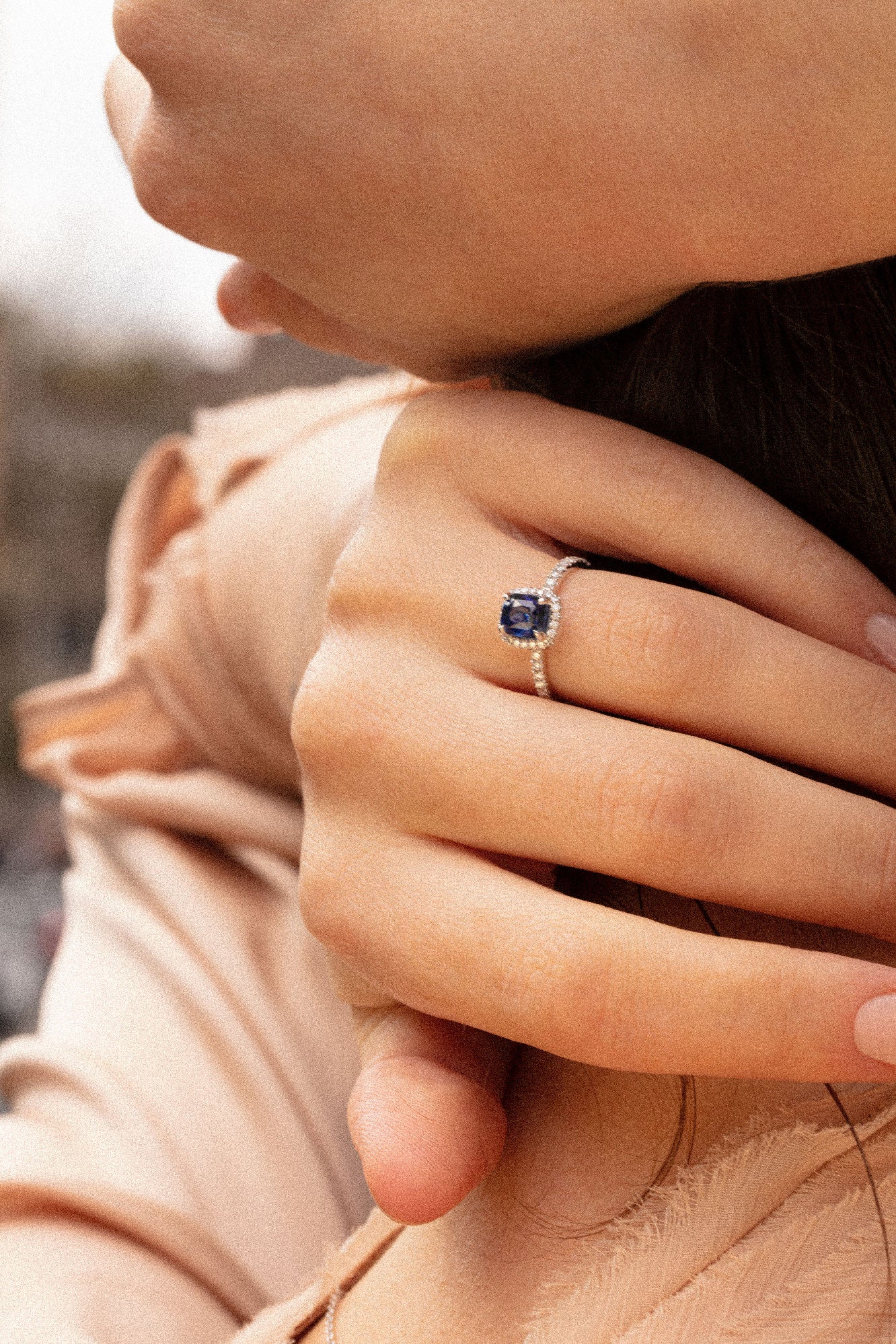 Photo d'une femme se tenant la nuque. Elle porte à son doigt une splendide bague en saphir bleu orné d'un halo en diamants blancs. La monture en or blanc 18 carats est pavée de scintillants diamants