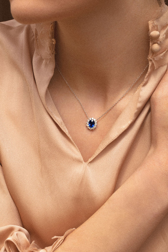 Photo d'une femme qui porte à son cou un collier en or blanc d'une grâce intemporelle. Il orne en son centre un ravissant saphir bleu. Le halo de diamants qui l'entoure magnifie les reflets bleus profond du saphir.